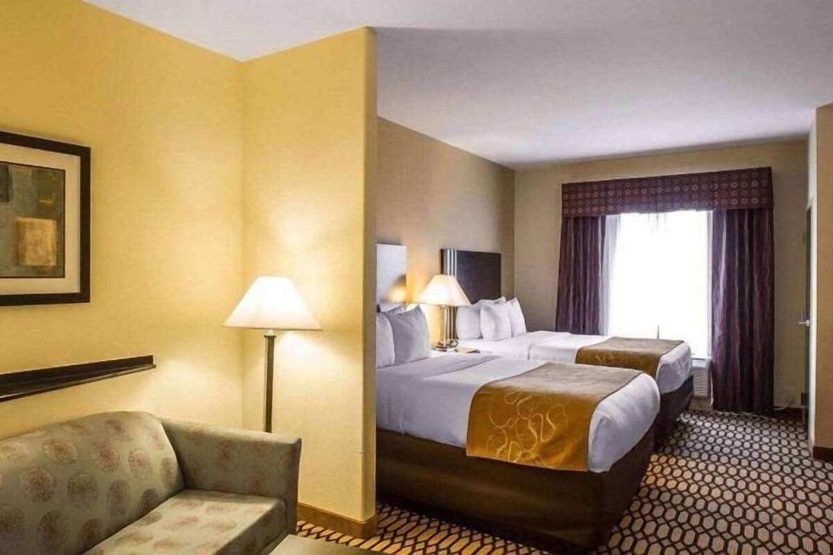 A hotel room with a sofa, two beds, a lamp, a painting, and a patterned floor.