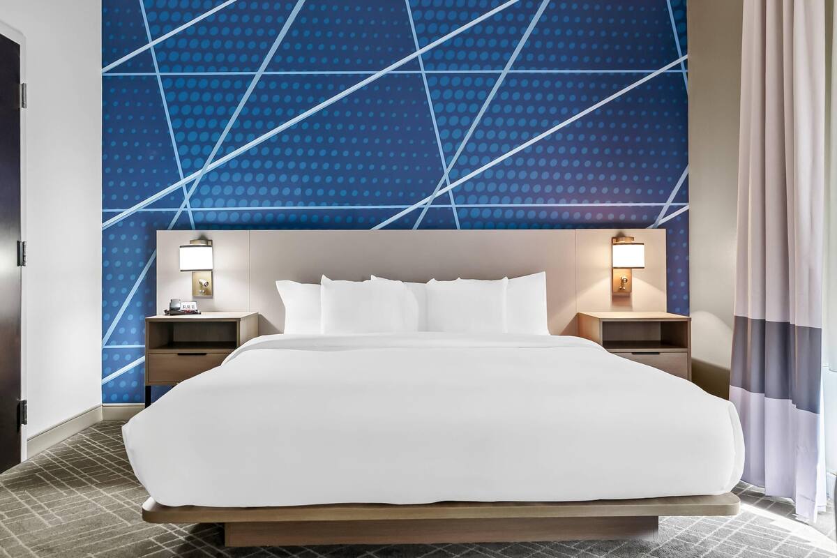 A hotel room with a large bed, two bedside tables with lamps, a wall with a geometric pattern, and a window with curtains.