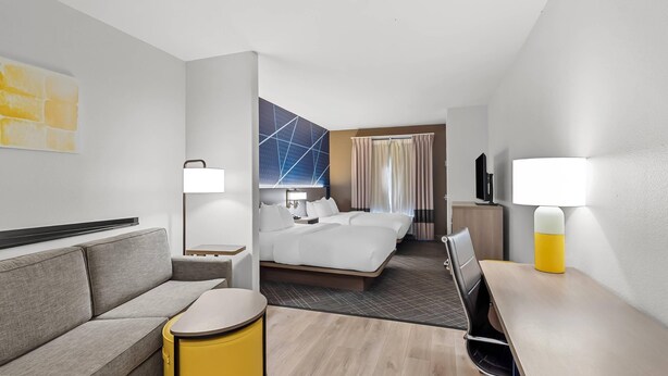 A hotel room with a sofa, a small round table, a bed with two pillows, a nightstand, a lamp, a TV, and a curtain. - Comfort Suites Simpsonville - Greenville (Simpsonville)
