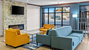 A modern living room with a stone wall, a flat-screen TV, a fireplace, and large windows with a view of the street. - Comfort Suites Simpsonville - Greenville (Simpsonville)