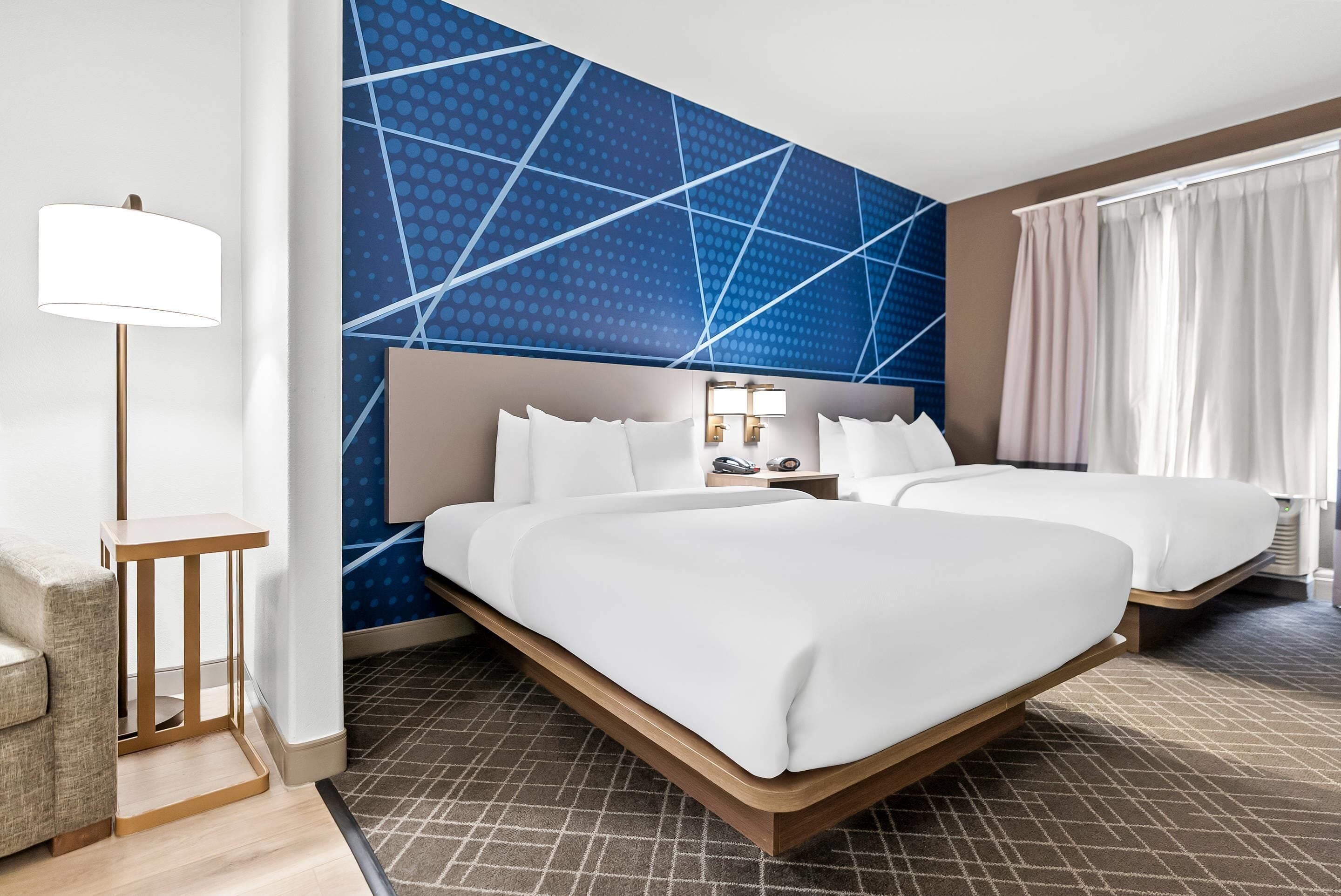 A hotel room with two beds, a desk, a lamp, and a patterned wall.
