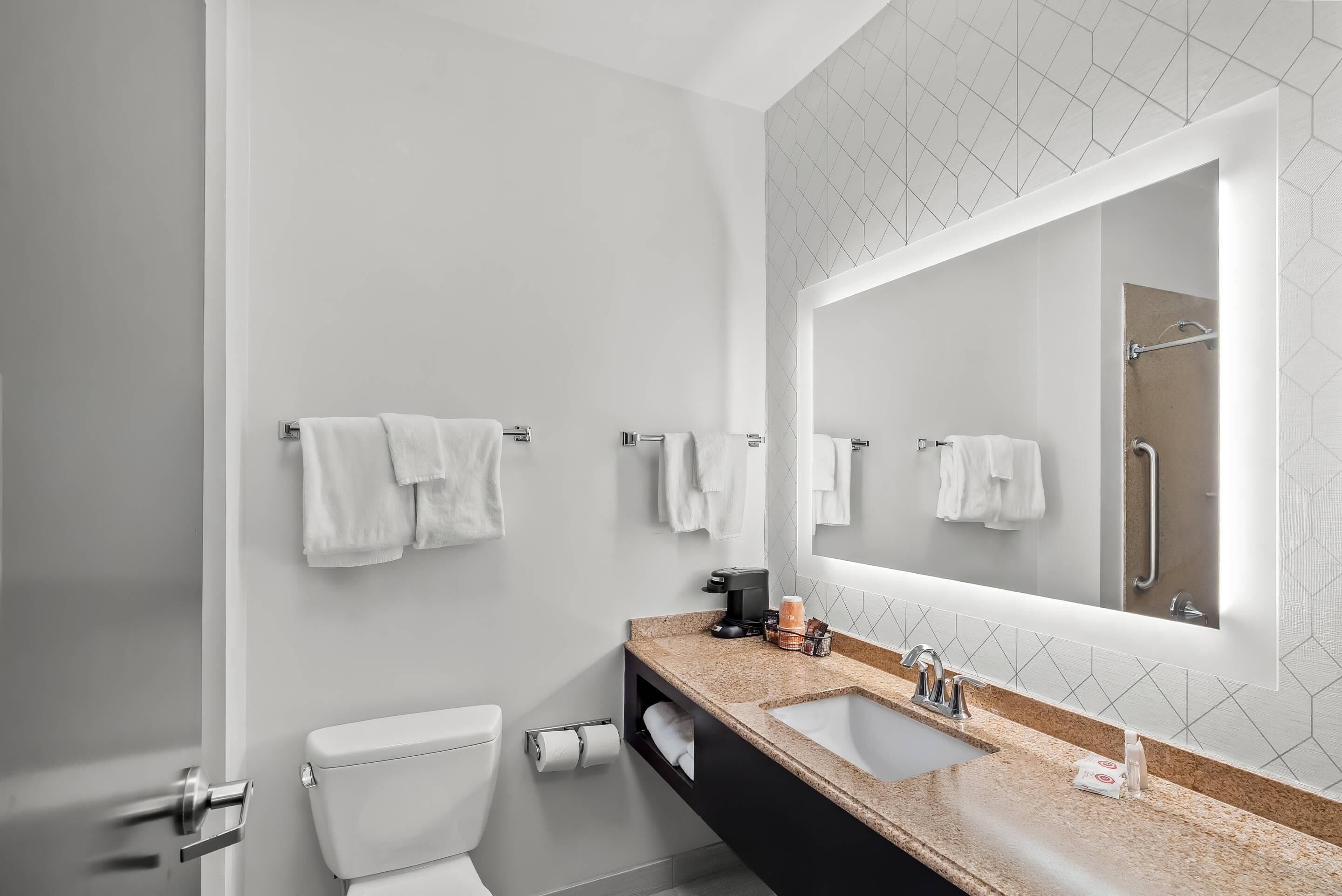 Un baño moderno con inodoro, lavamanos y espejo. Hay toallas colgadas en la pared y se ve una zona de ducha en el reflejo del espejo.