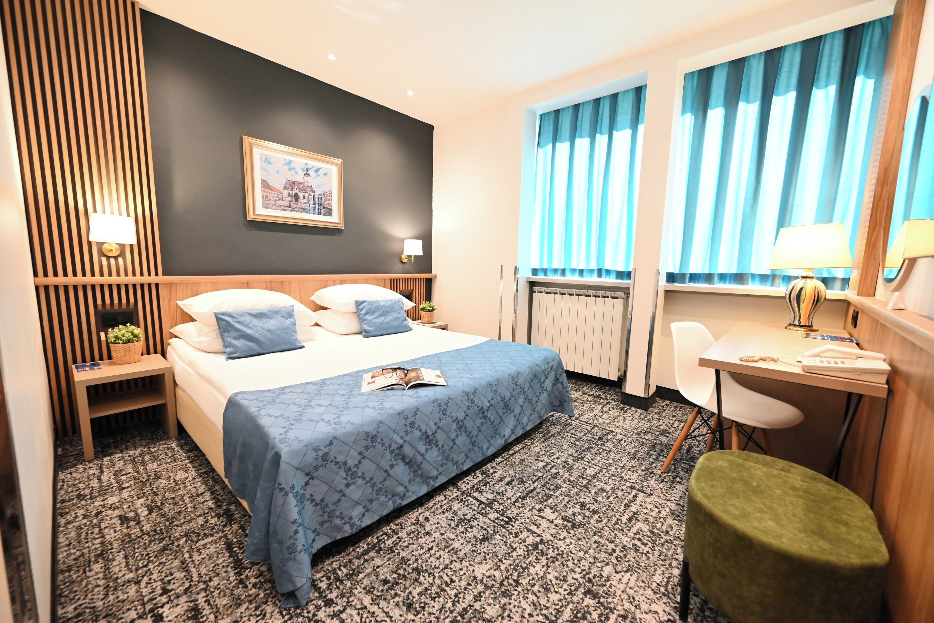 Una habitación de hotel con cama, mesitas de noche, escritorio, silla y un taburete pequeño.