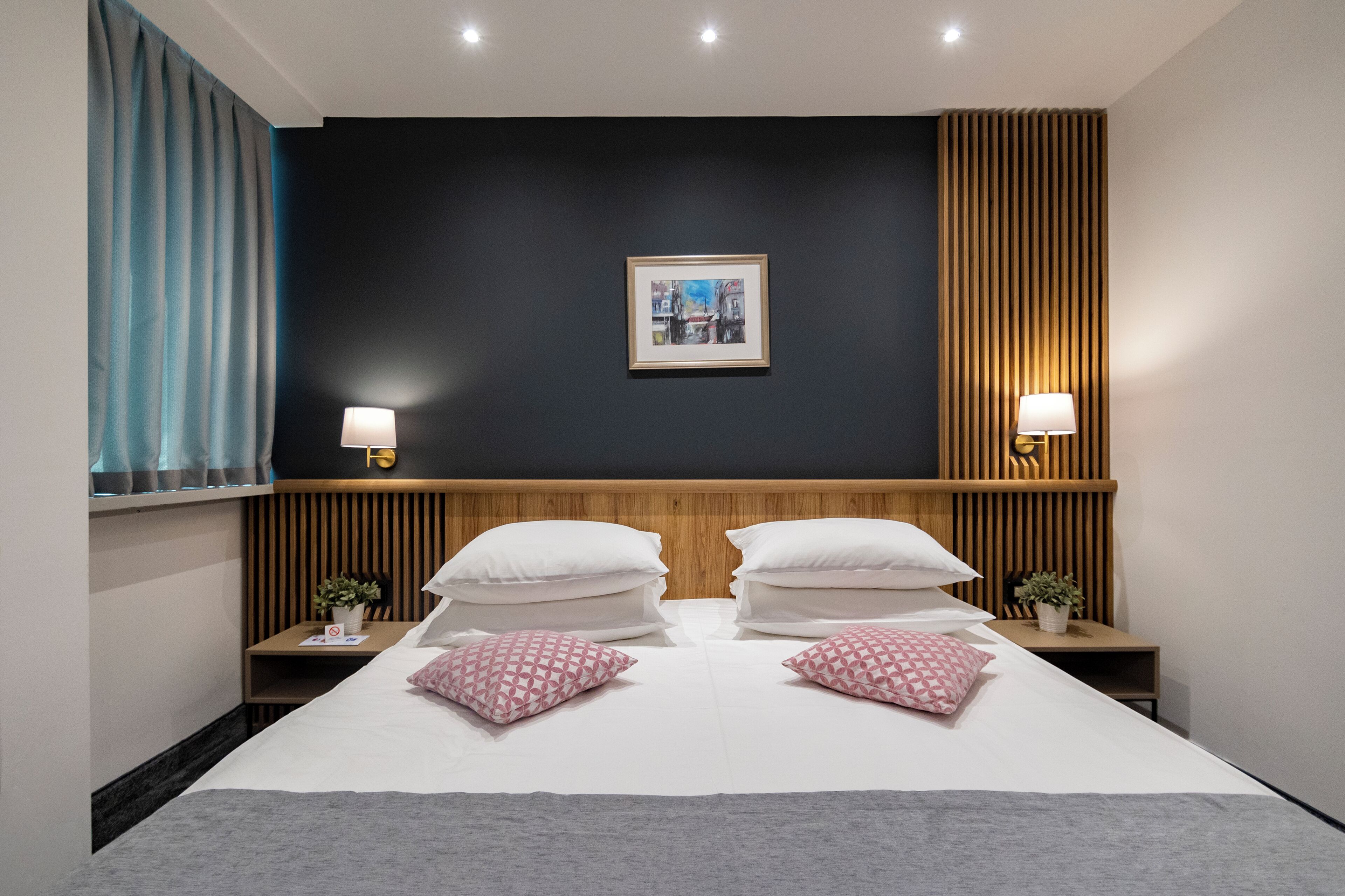 A hotel room with a large bed, two bedside tables, a nightstand, a wall-mounted lamp, a framed picture, and a window with curtains.