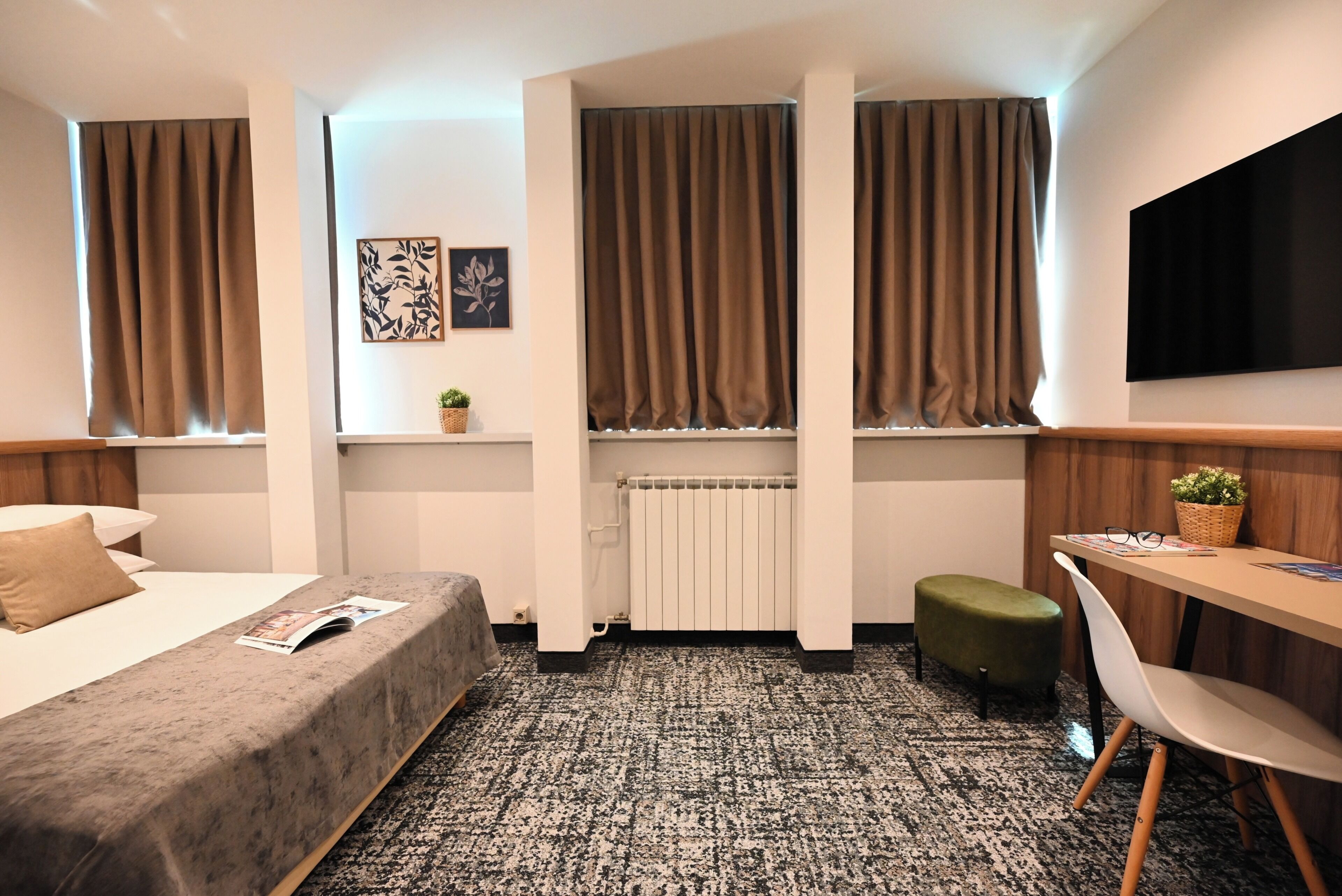 Habitación de hotel con cama, escritorio, silla, radiador y ventana con cortinas.