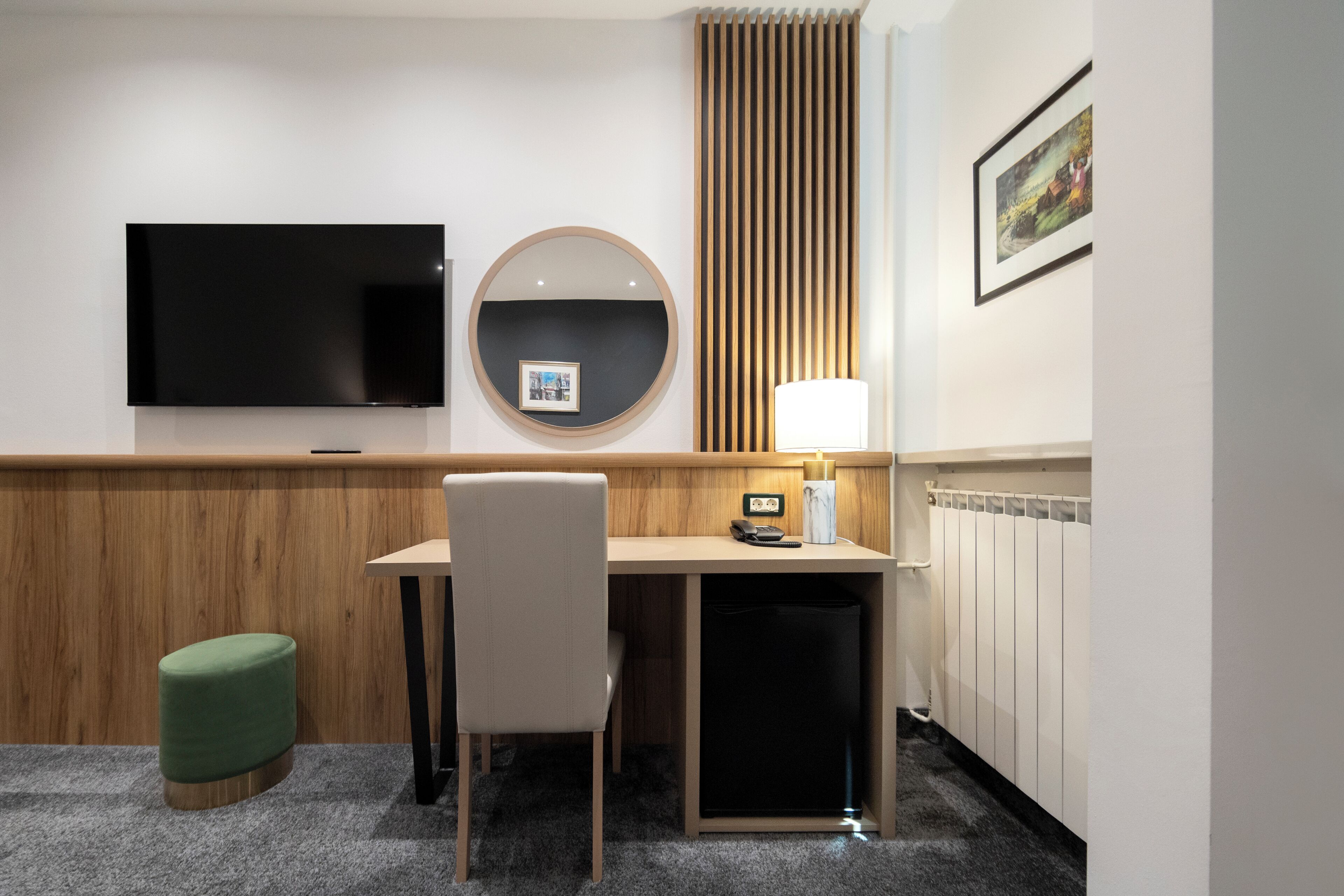 A hotel room with a flat-screen TV, a round mirror, a desk with a lamp, a telephone, and a green ottoman.