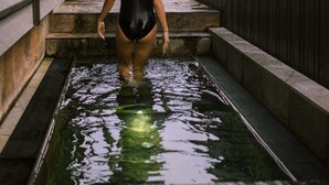 Una persona con traje de baño negro caminando por una piscina con escalones de piedra y una valla de madera al fondo.