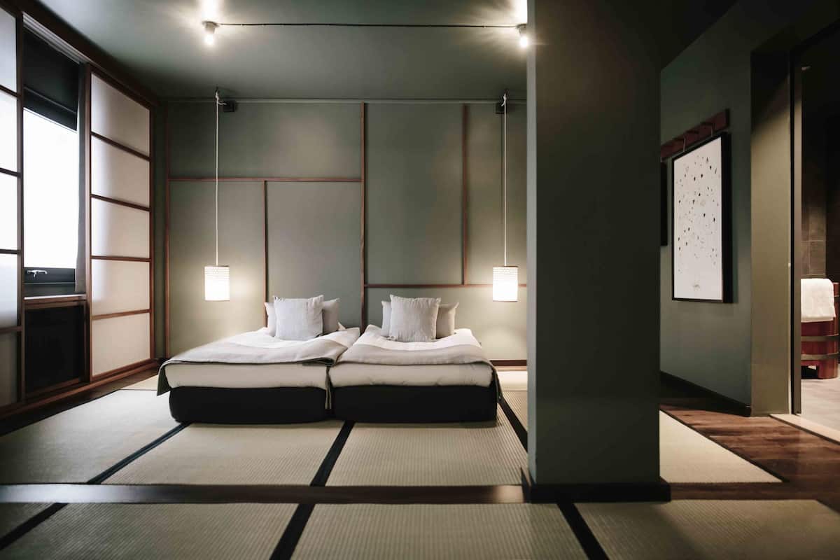 Una habitación tradicional japonesa con suelo de tatami, una cama baja con cabecera oscura y una lámpara colgante.