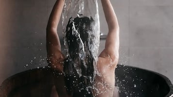 A person in a wooden bathtub filled with water, holding a bowl above their head.