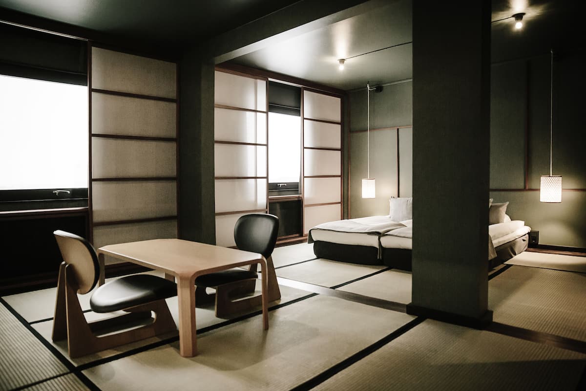 Una habitación tradicional japonesa con suelo de tatami, una mesa baja con una silla, una cama y un puertas correderas.
