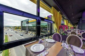 A restaurant with tables set for dining, large windows offering a view of a road and buildings, and yellow curtains on the windows. - Business V Hotel (Tirgu Mures)