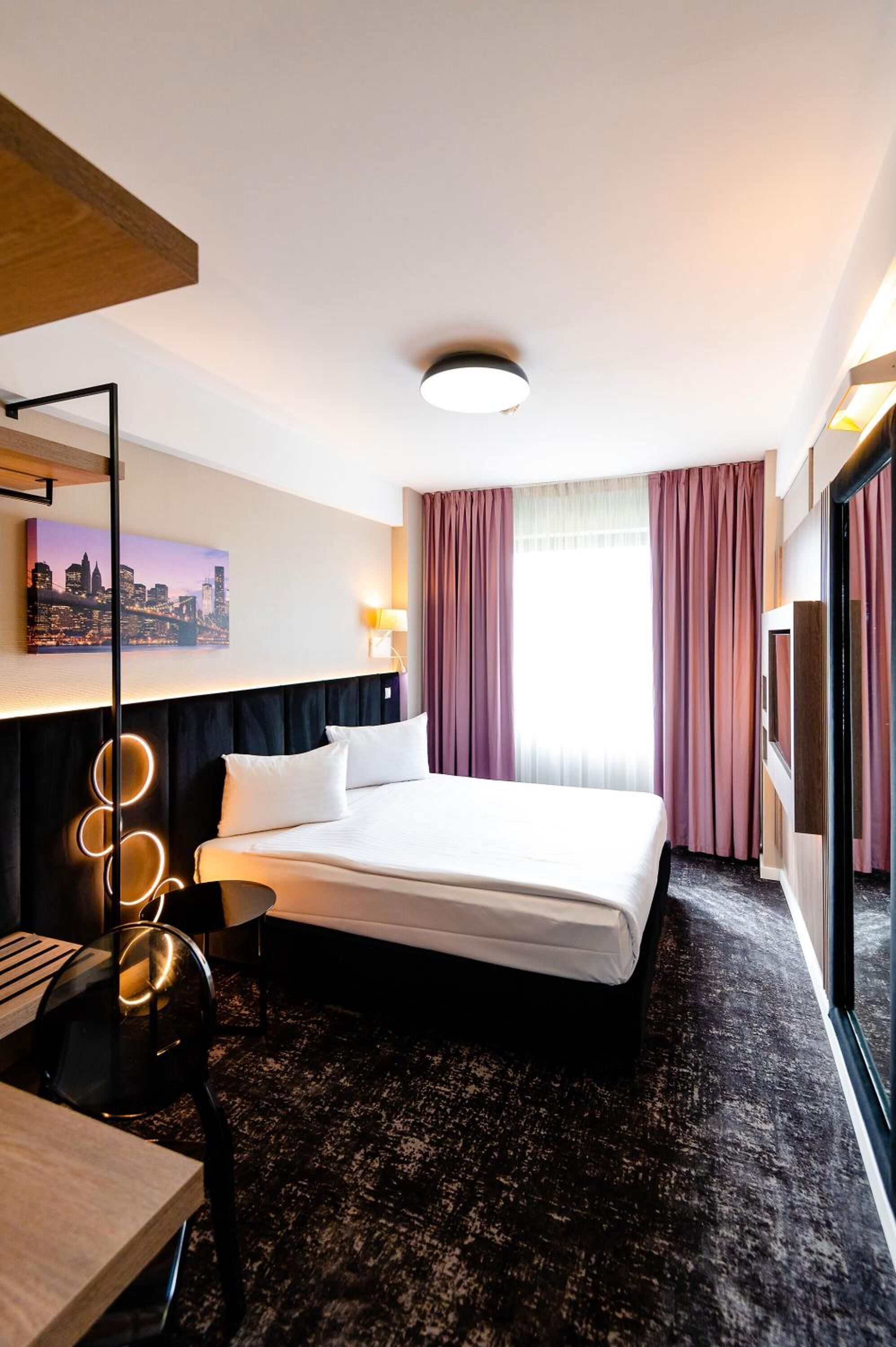 A hotel room with a bed, a desk, a chair, a cityscape painting, a round mirror, and a large window with curtains.
