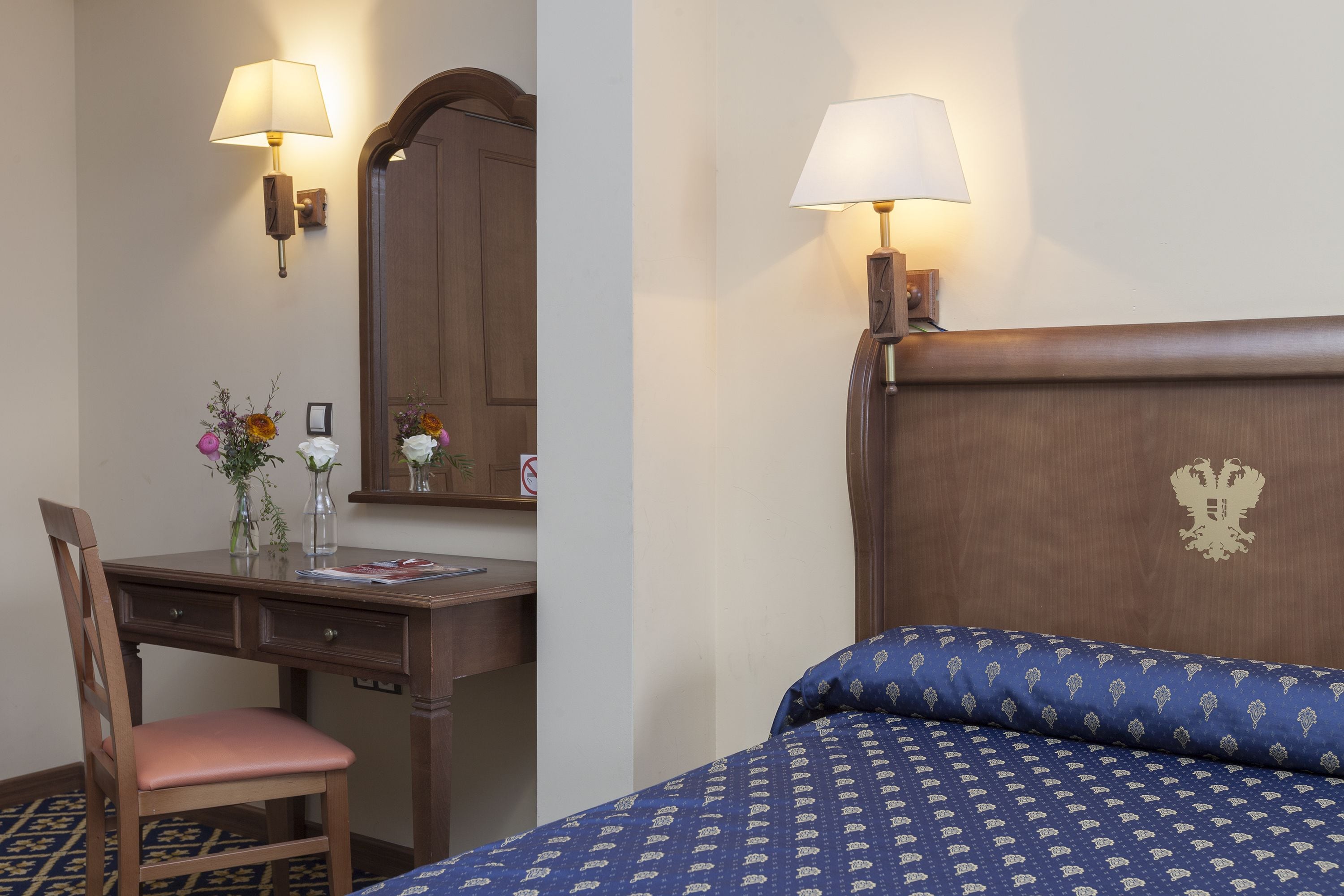 Een hotelkamer met een houten bureau, een stoel, een bed met een blauw beddegoed met patroon, en twee wandlampen.