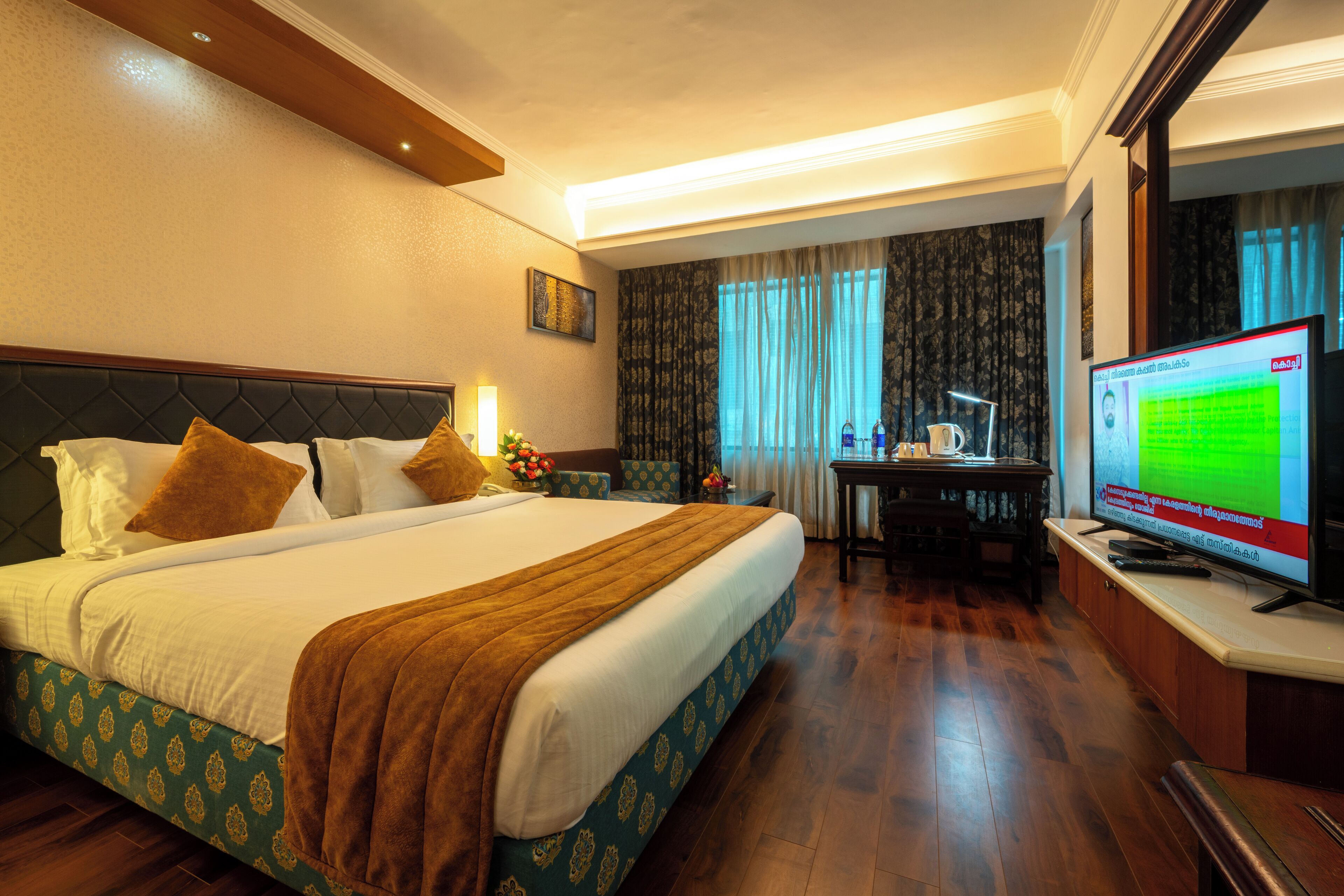 A hotel room with a large bed, a desk with a computer, a television, and a window with curtains.