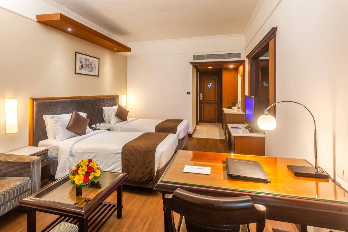 A hotel room with two beds, a desk, a chair, a lamp, and a vase of flowers.