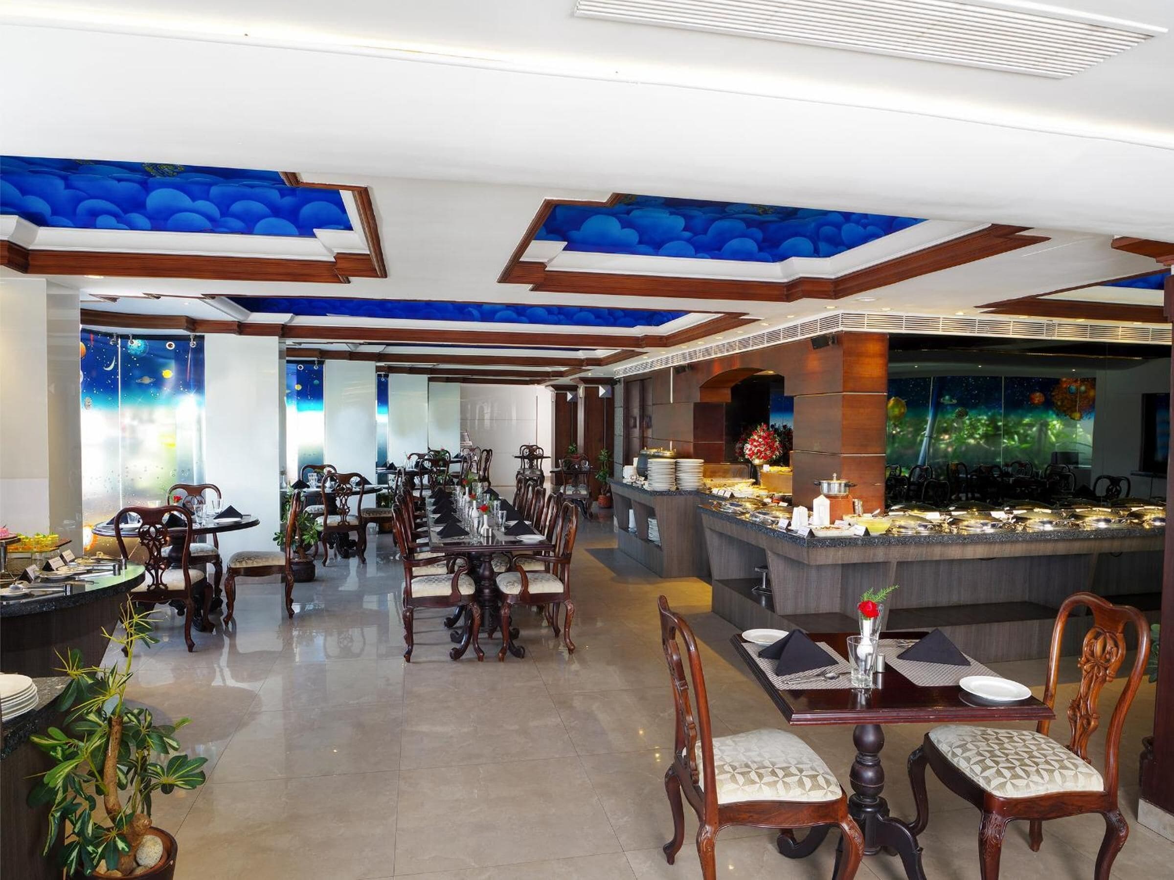 A spacious dining area with multiple tables and chairs, a buffet counter, and a ceiling with a blue sky design.