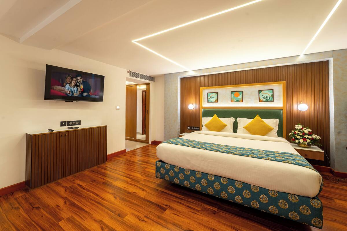 A hotel room with a large bed, a flat-screen TV mounted on the wall, a wooden headboard, and a wooden floor.