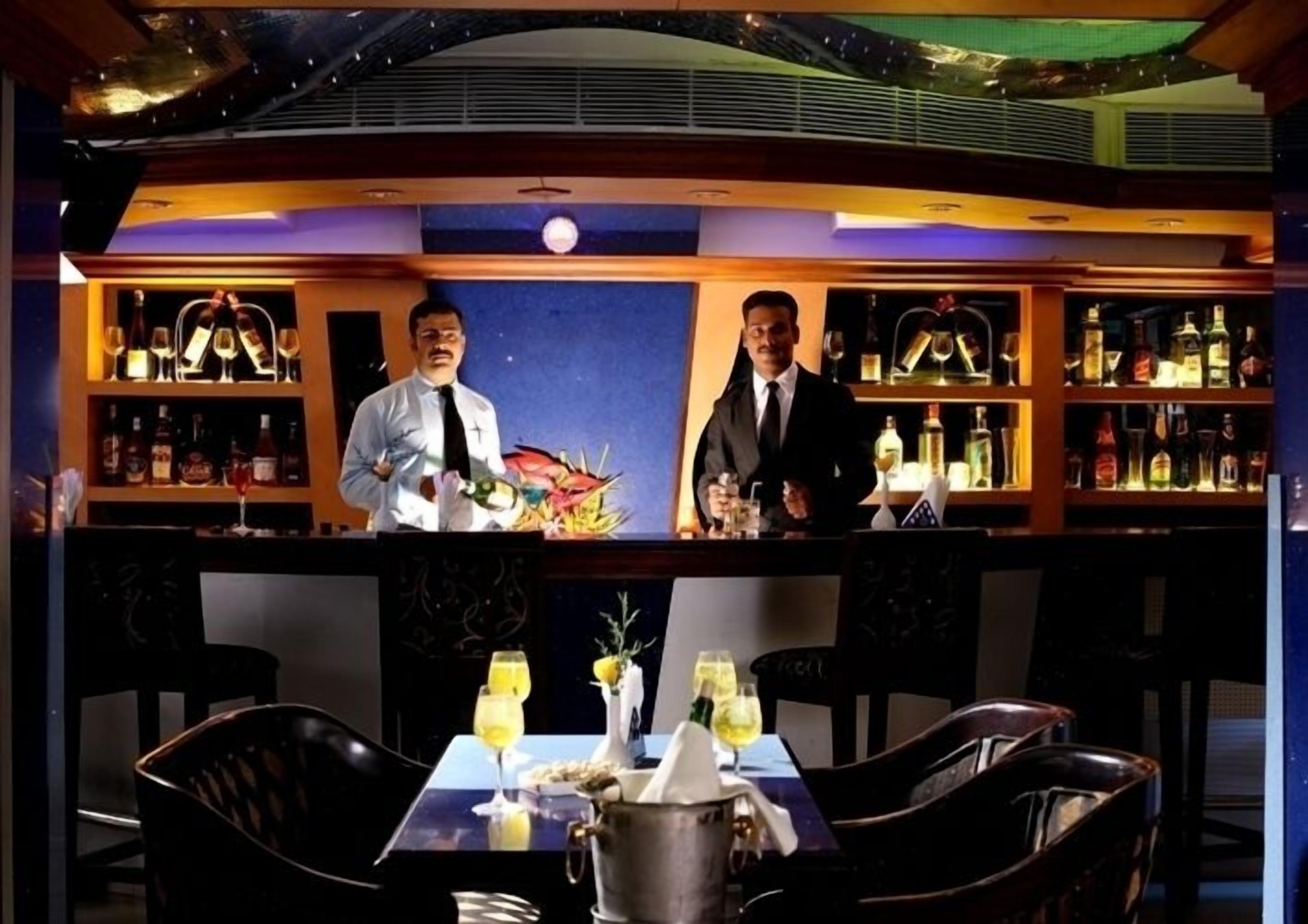A bar area with two bartenders, a bar counter, shelves with bottles, and a table with chairs and a vase of flowers.