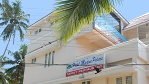 A two-story building with a sign for 'Hotel Marine Palace' and 'Family Restaurant'. There are flags in front of the building, a parked car, and a security guard. - Hotel Marine Palace (Neyyattinkara)