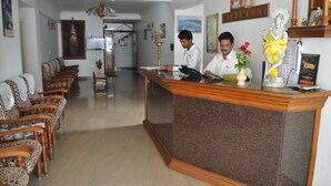 A reception area with a reception desk, two men at the desk, a sofa with patterned cushions, and a staircase in the background. - Hotel Marine Palace (Neyyattinkara)