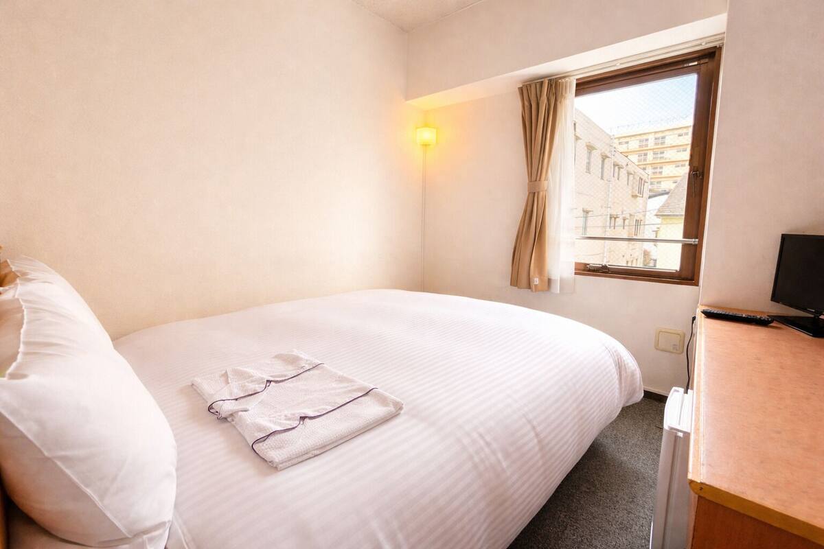 A hotel room with a bed, a desk with a computer, a window with curtains, and a wall-mounted light.