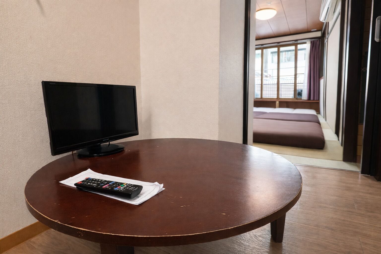 A round wooden table with a remote control and papers, a flat-screen TV, and a view of a room with a bed and curtains.