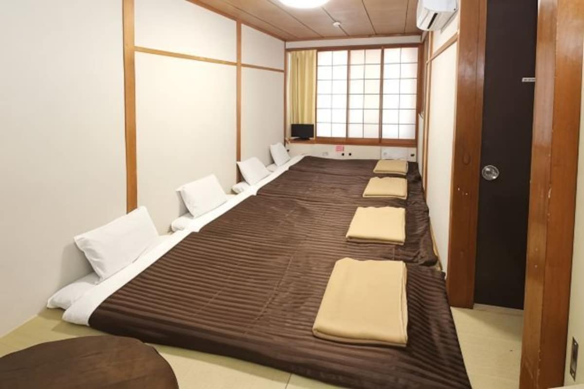 A hotel room with a long bed, multiple pillows, a wooden floor, a window with curtains, and a door.