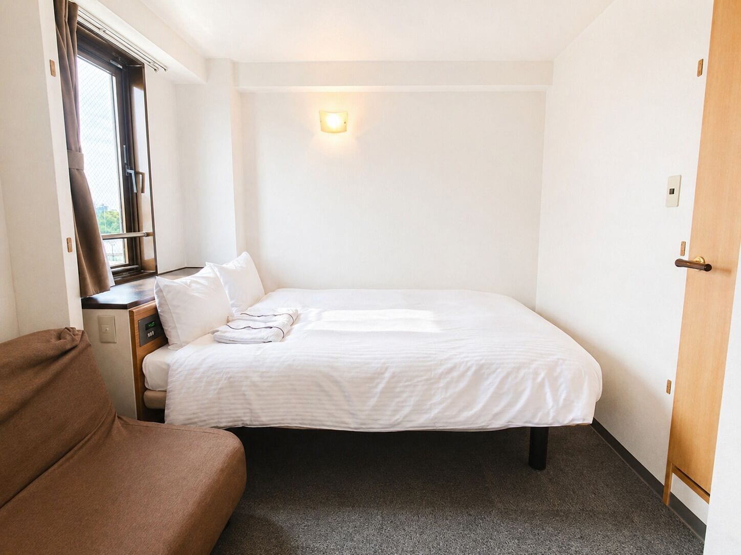 A single bed with white bedding, a bedside table, a window with curtains, and a door.