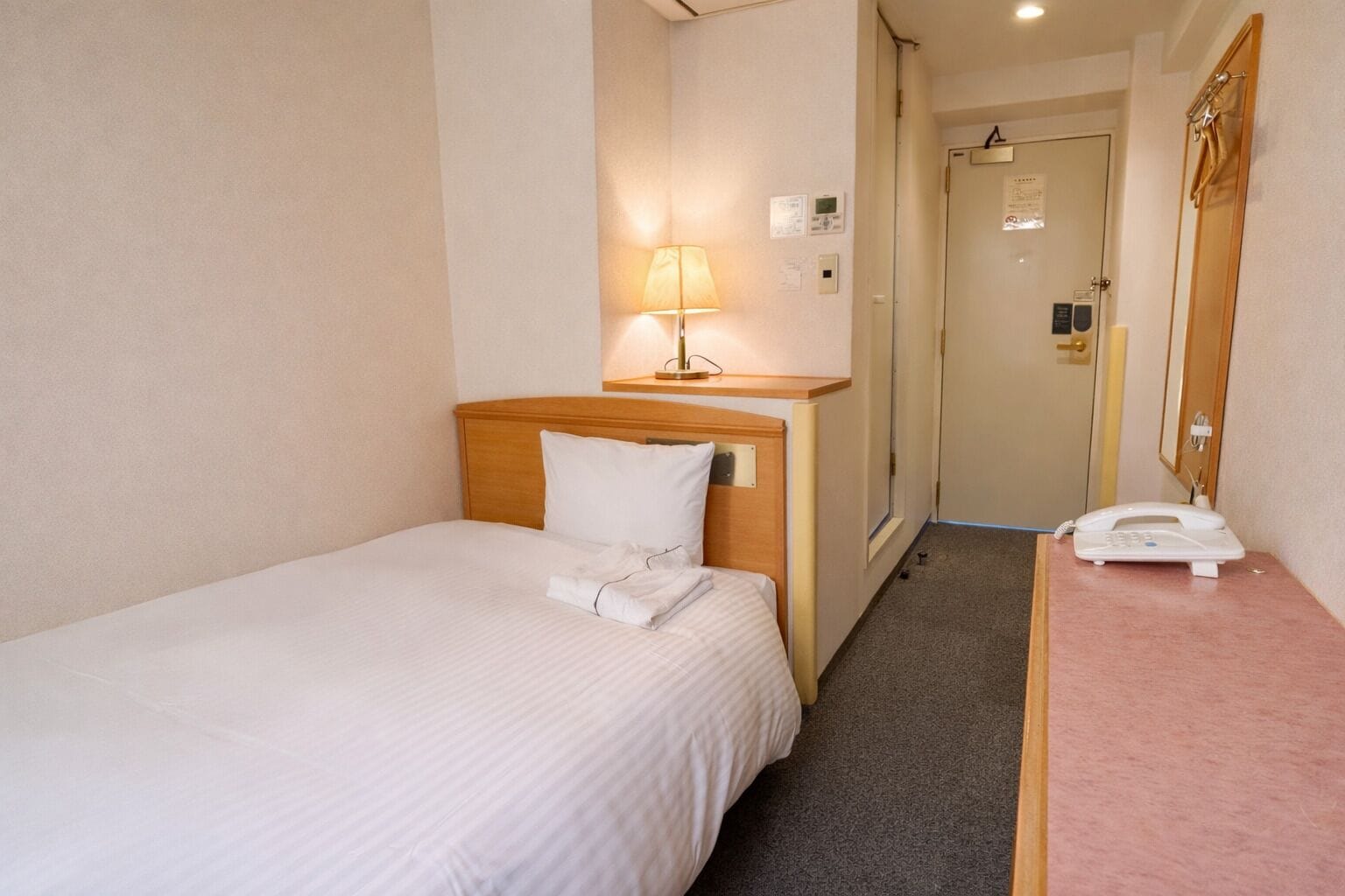A hotel room with a single bed, a bedside lamp, a telephone, and a door leading to a bathroom.