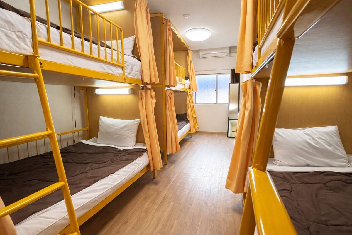 A dormitory room with bunk beds, a wooden floor, and a window with curtains.