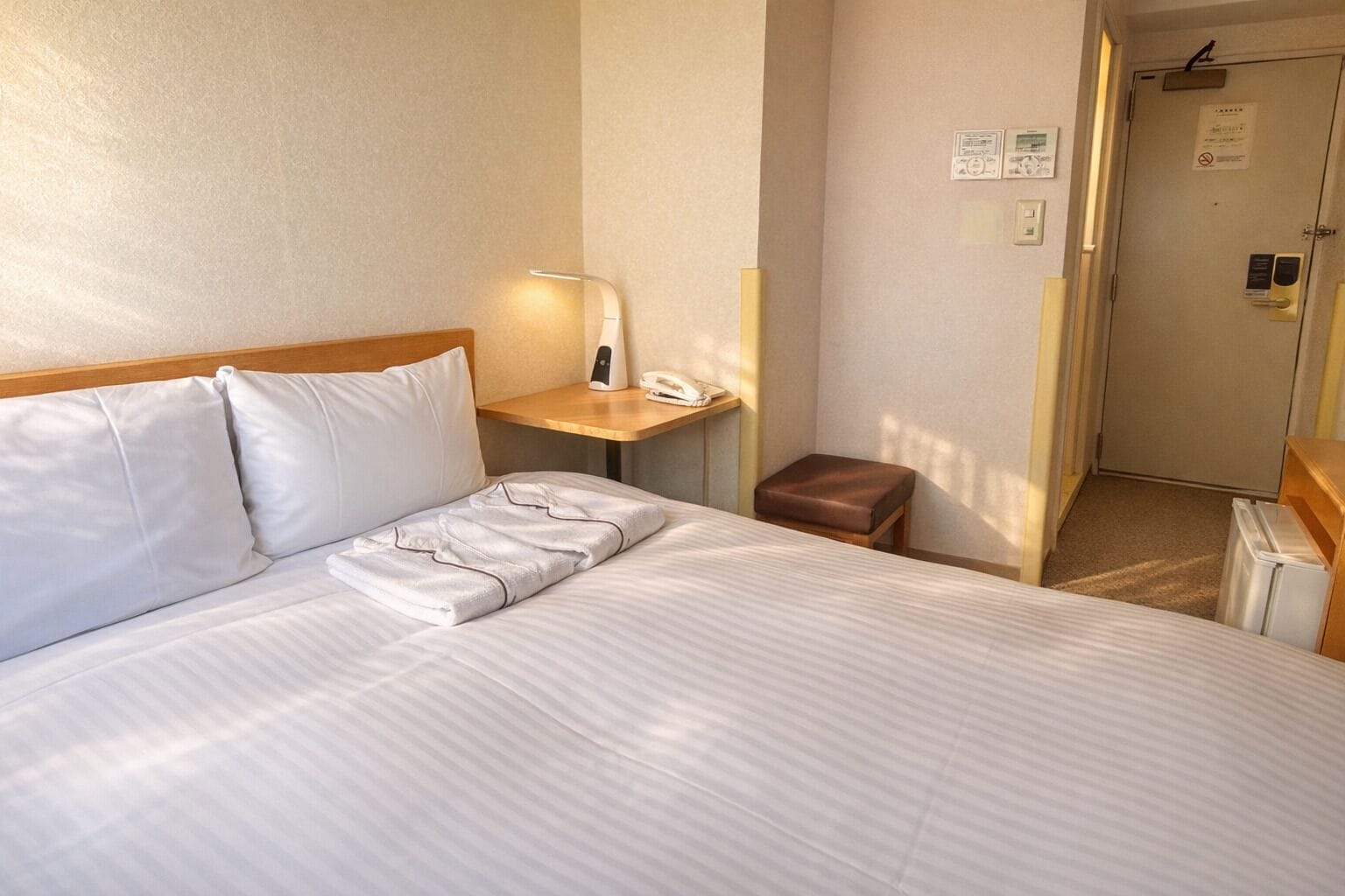 A hotel room with a bed, a bedside table with a lamp, a telephone, a brown bench, and a door with a sign.
