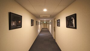 A long, narrow hallway with framed pictures on the walls, a carpeted floor, and a ceiling with recessed lighting. - Sakura Hotel Hatagaya (Tokyo)