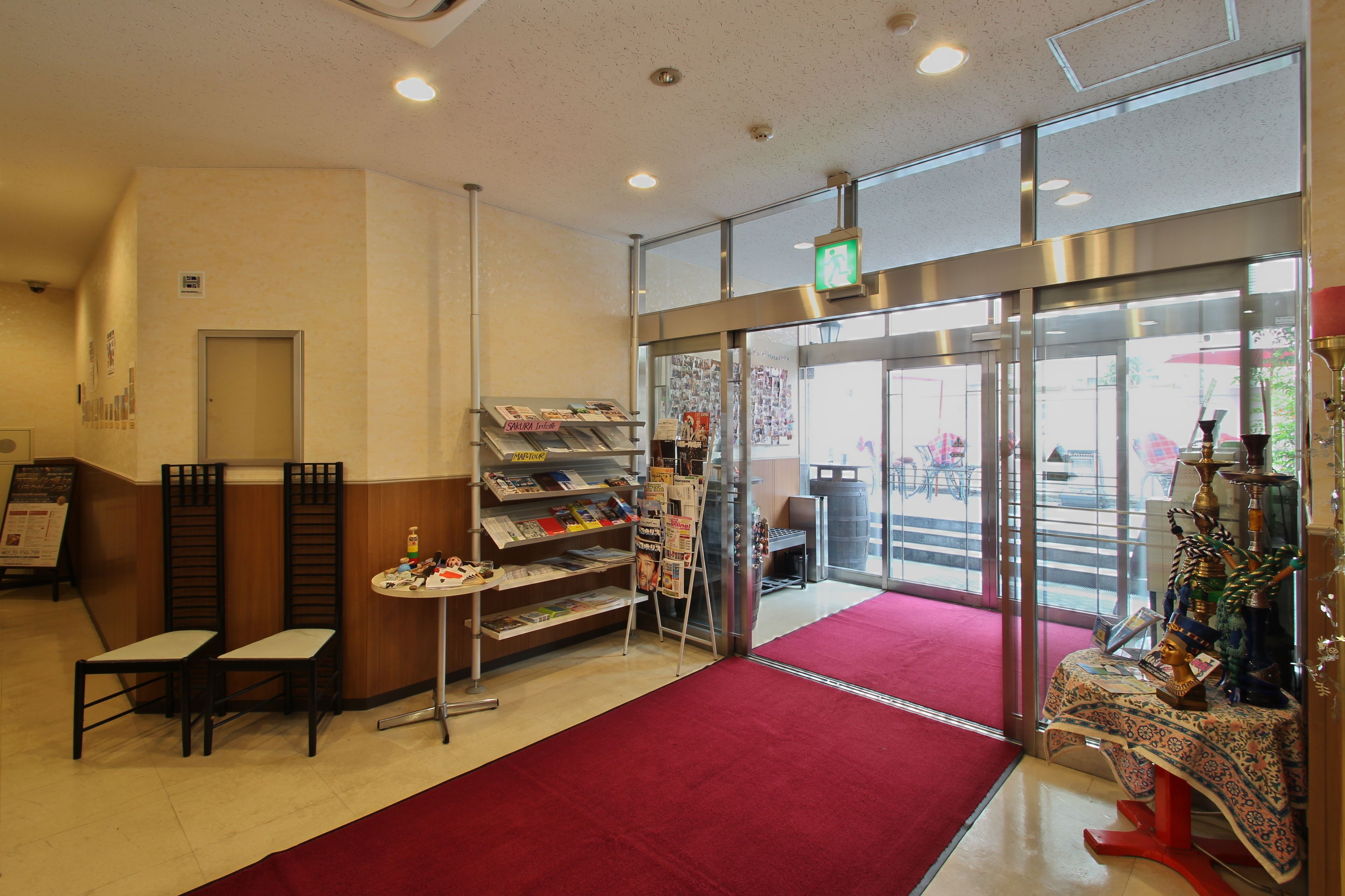 A hotel lobby with a red carpet, a reception desk, a glass door entrance, and a small table with decorative items.