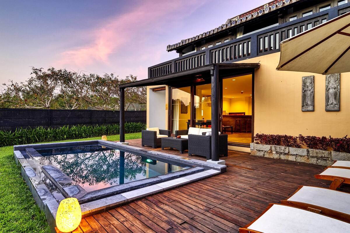 A modern outdoor pool area with a wooden deck, lounge seating, and a covered patio.