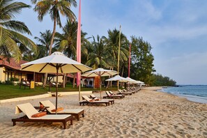 Ein Strand mit Reihen von Liegestühlen und Sonnenschirmen, Palmen und einem Gebäude mit rotem Dach.
