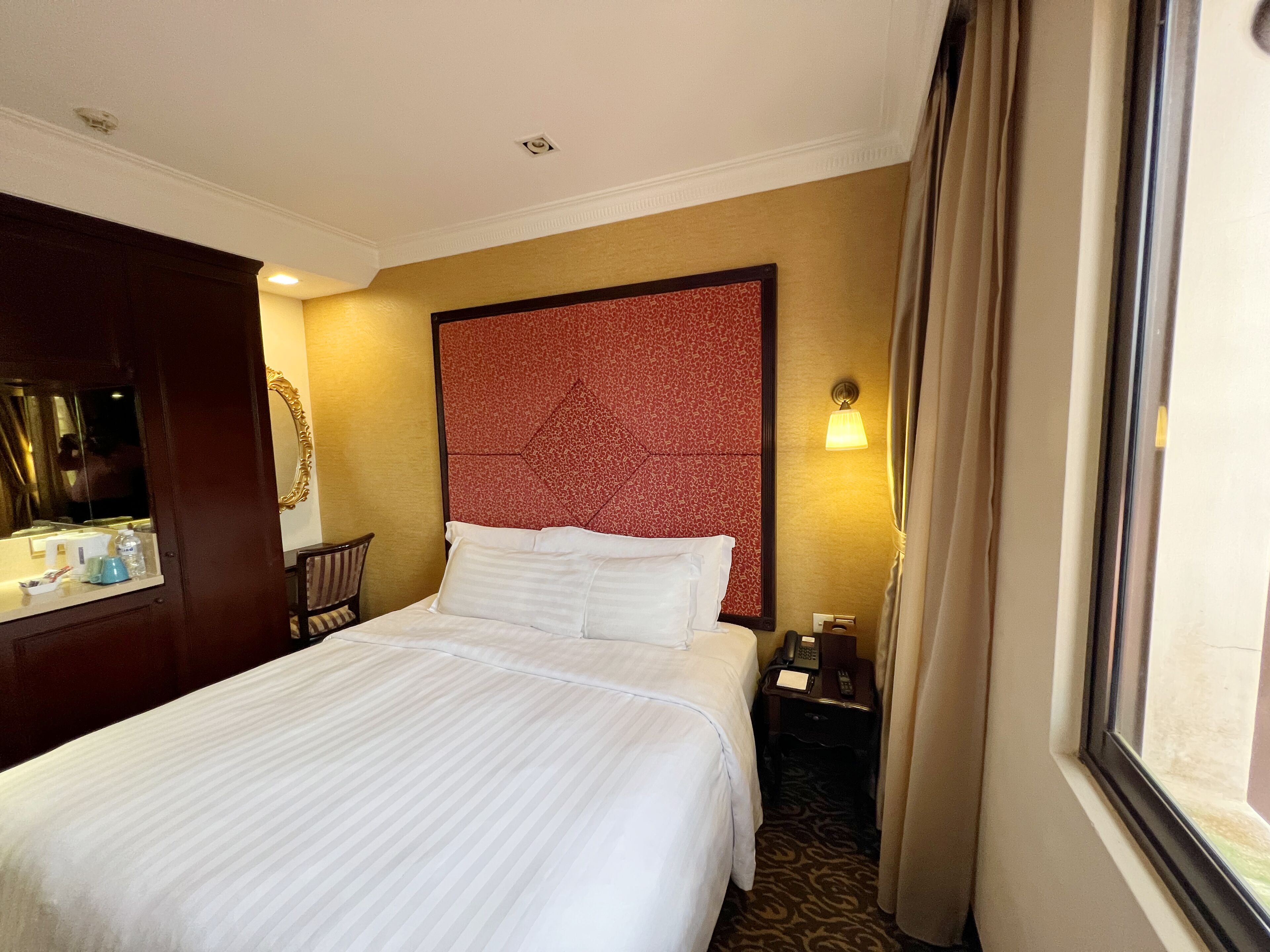 Een hotelkamer met een groot bed, een houten nachtkastje met telefoon, een spiegel en een raam met gordijnen.