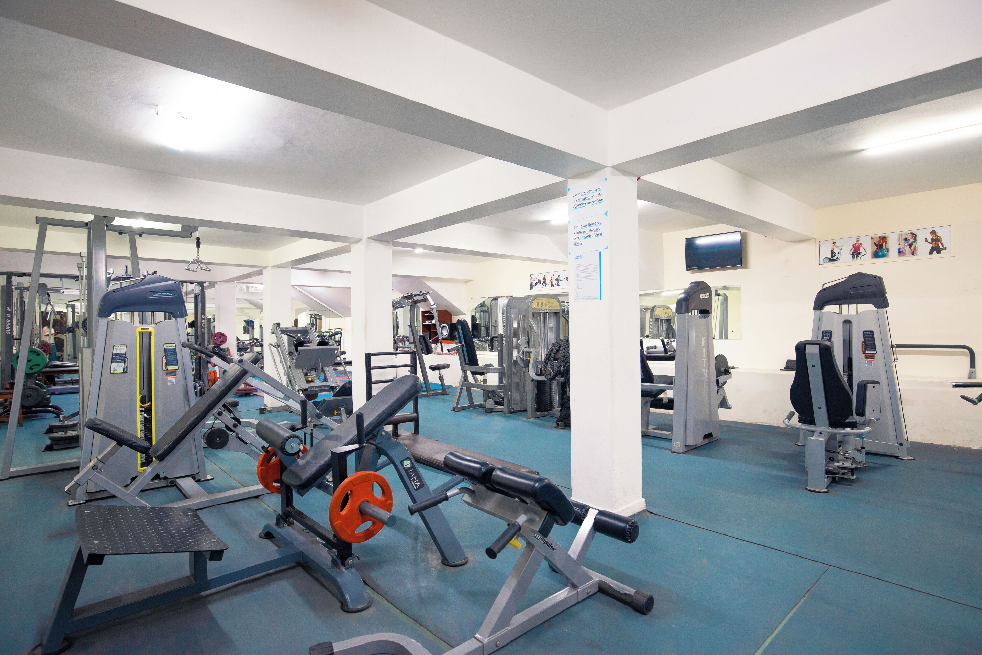 A gym with various exercise equipment, a television on the wall, and a poster with fitness illustrations.