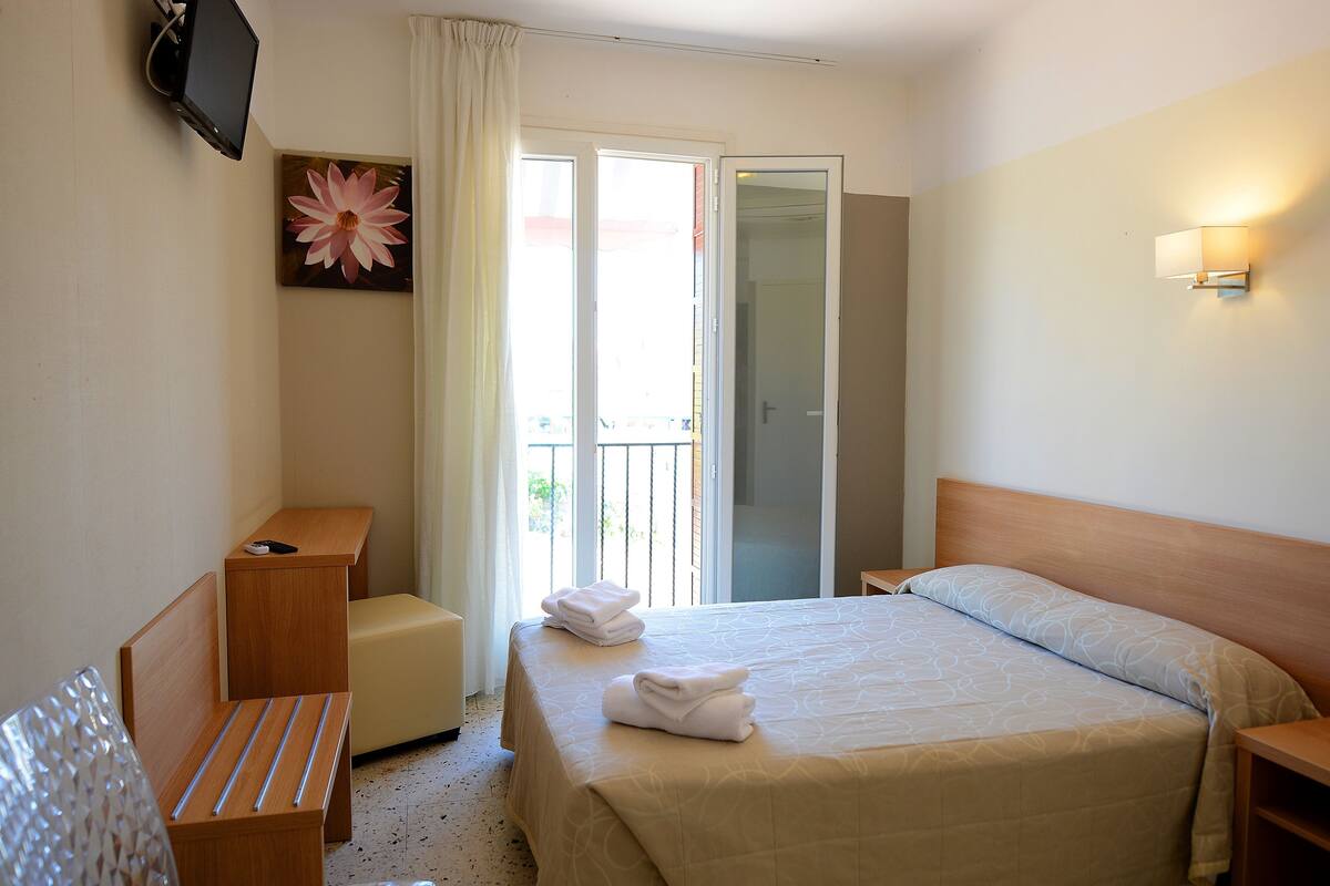 A hotel room with a bed, a desk, a chair, a TV, and a balcony with a view.
