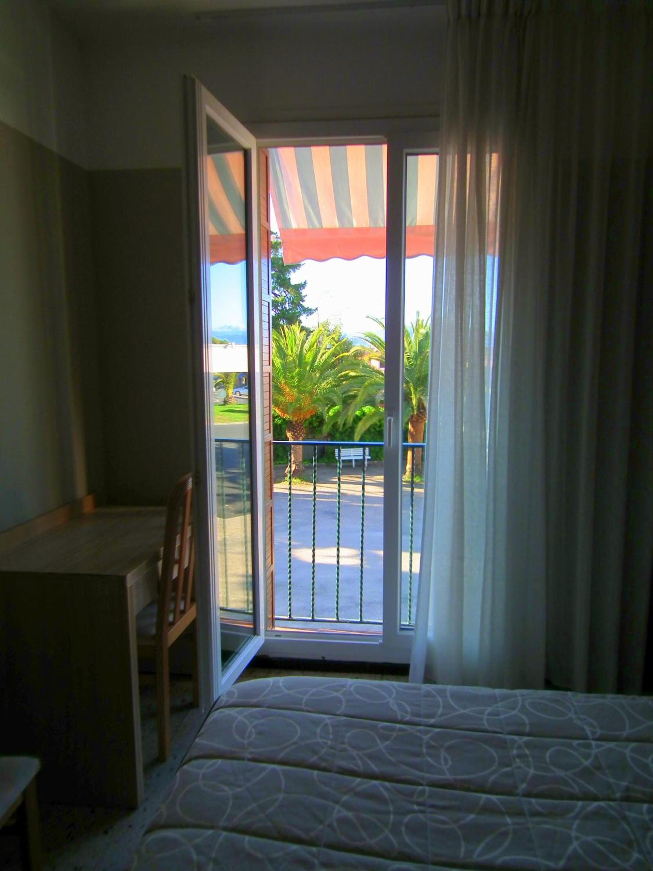 A room with a bed, a desk, and a chair. There is a sliding glass door leading to an outdoor area with plants and a fence.