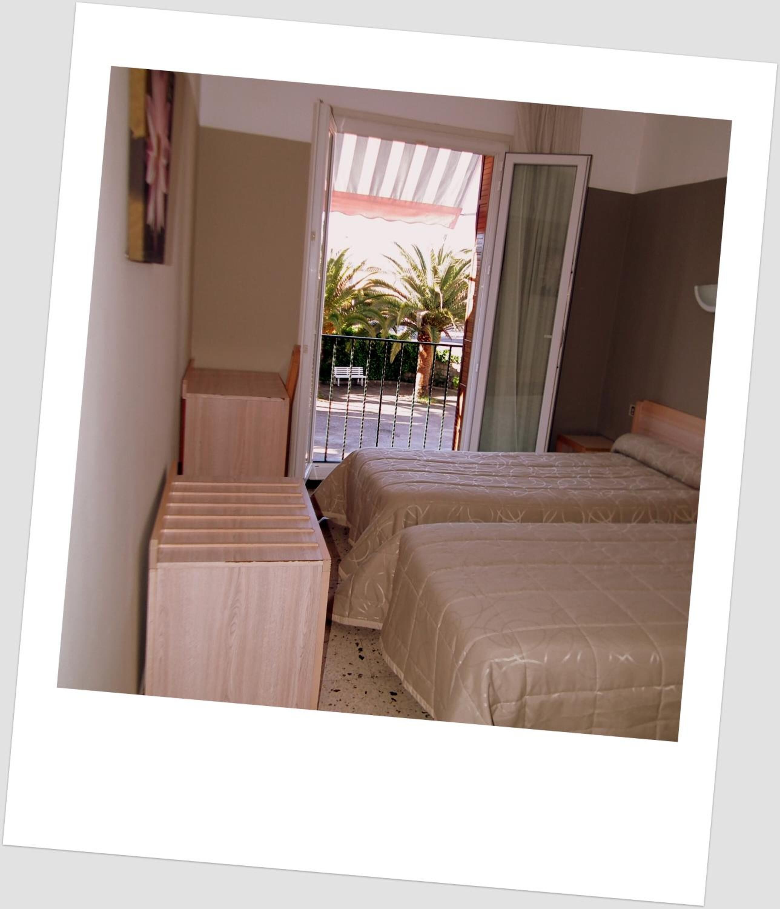 A bedroom with two beds, a wooden bench, a window with a view of palm trees, and a balcony.