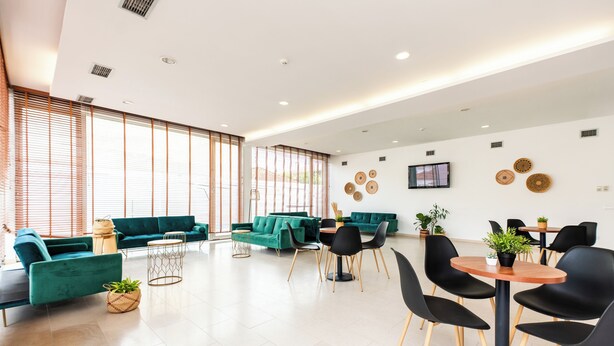 A spacious room with teal sofas, a dining area with black chairs, and a large window with blinds. - Hotel Evenia Monte Real (Leiria)
