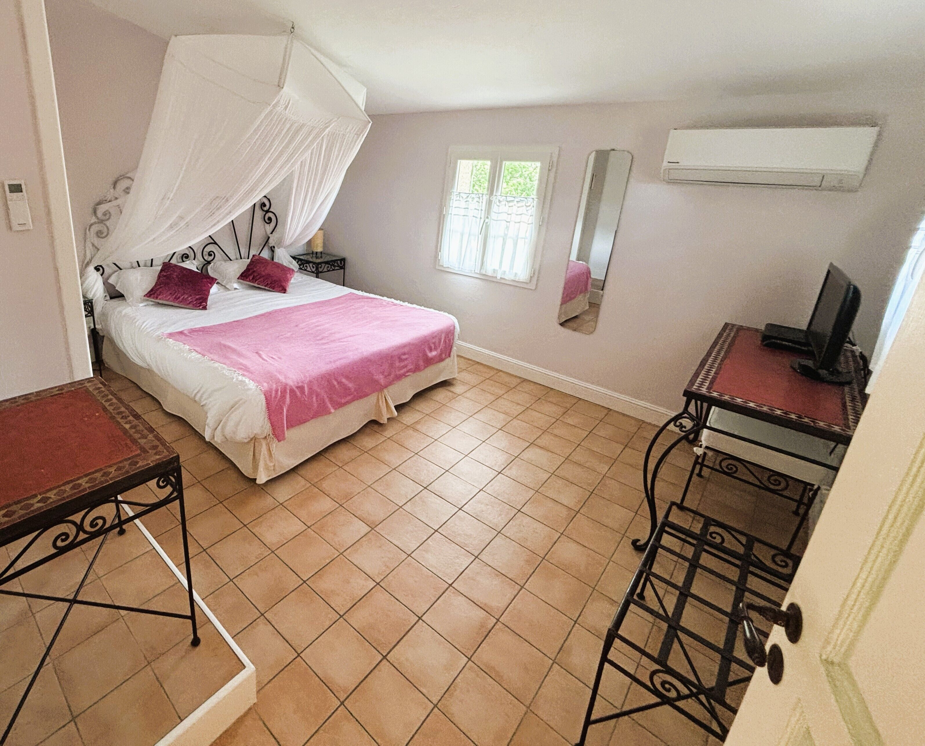 A bedroom with a bed, a bedside table, a chair, a window, and an air conditioning unit.