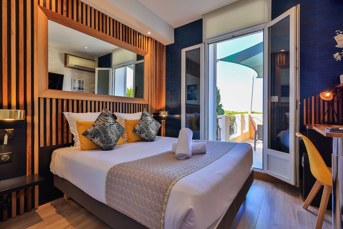 A modern hotel room with a large bed, a wooden headboard, a large mirror, and a sliding glass door leading to a balcony.