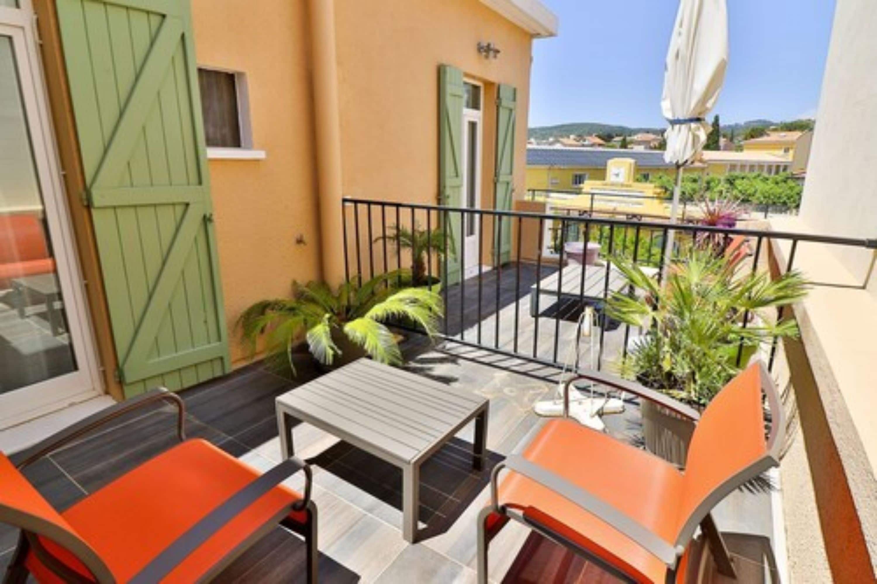 Un balcón con una mesa y sillas, una puerta con contraventanas verdes y vista al mar.