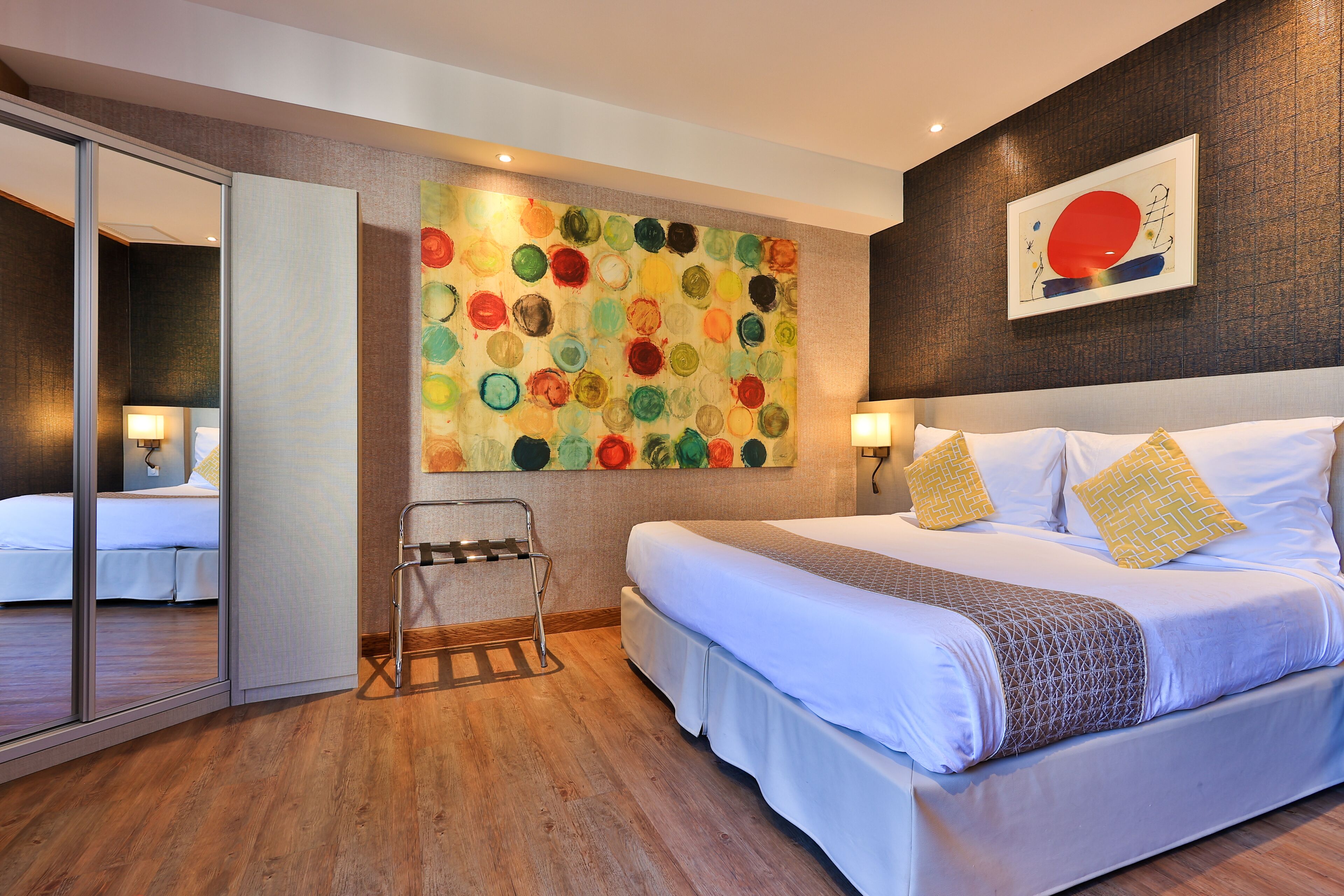 A modern hotel room with a large bed, a glass partition, and a colorful abstract painting on the wall.
