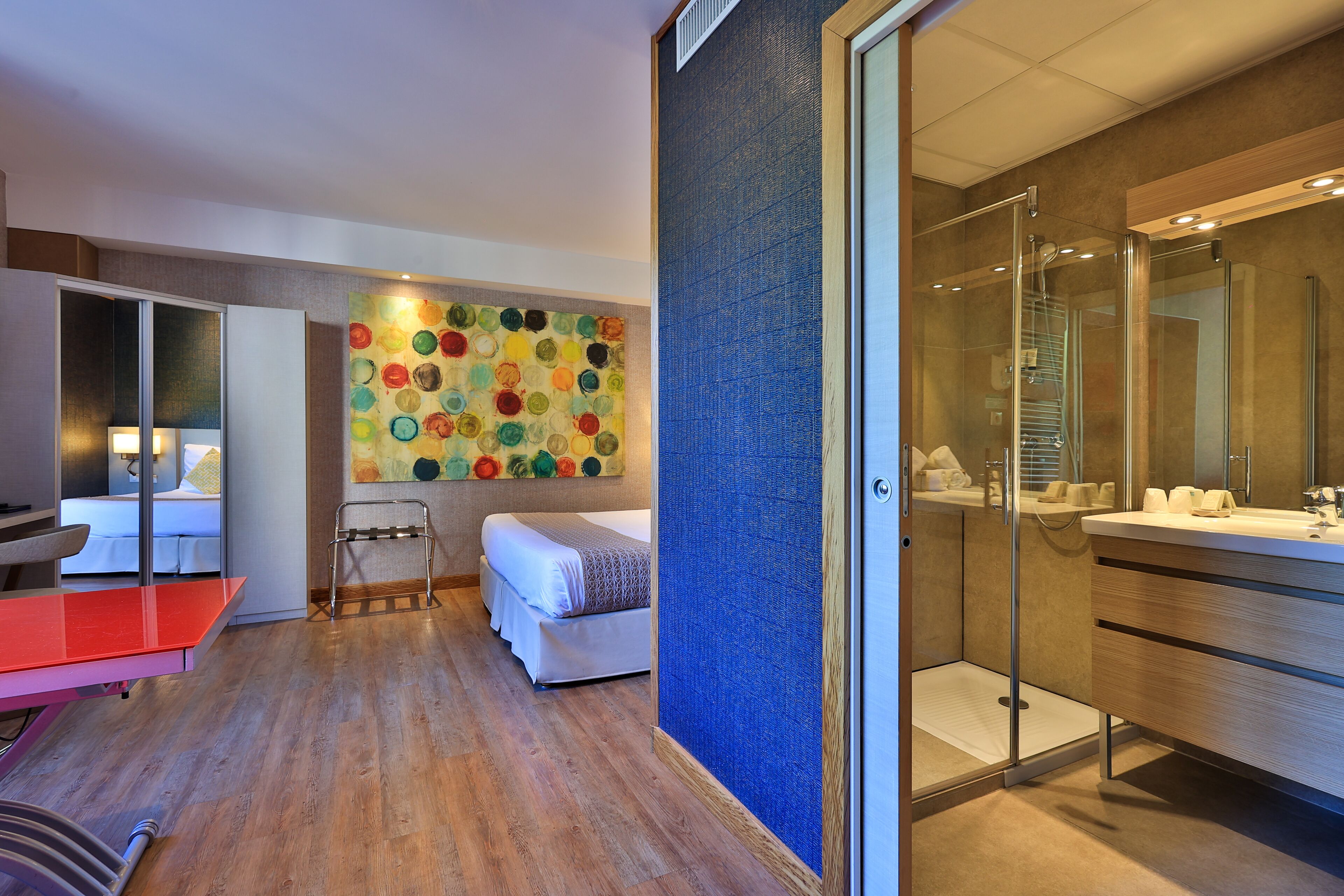 A modern hotel room with a wooden floor, a bed, a red table, a colorful wall art, a glass shower, and a large mirror.
