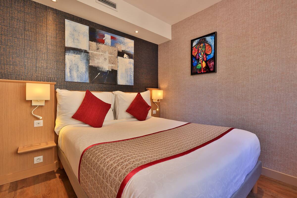 A hotel room with a large bed, two red pillows, a geometric patterned bedspread, a wall art piece, and a small table with a lamp.