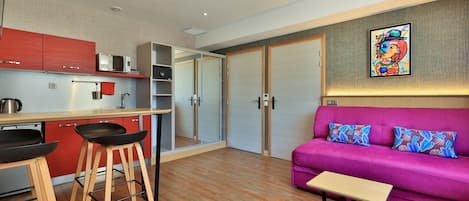 A modern kitchen with red cabinets, a microwave, and a coffee maker. There is a purple sofa with patterned cushions and a small wooden table.