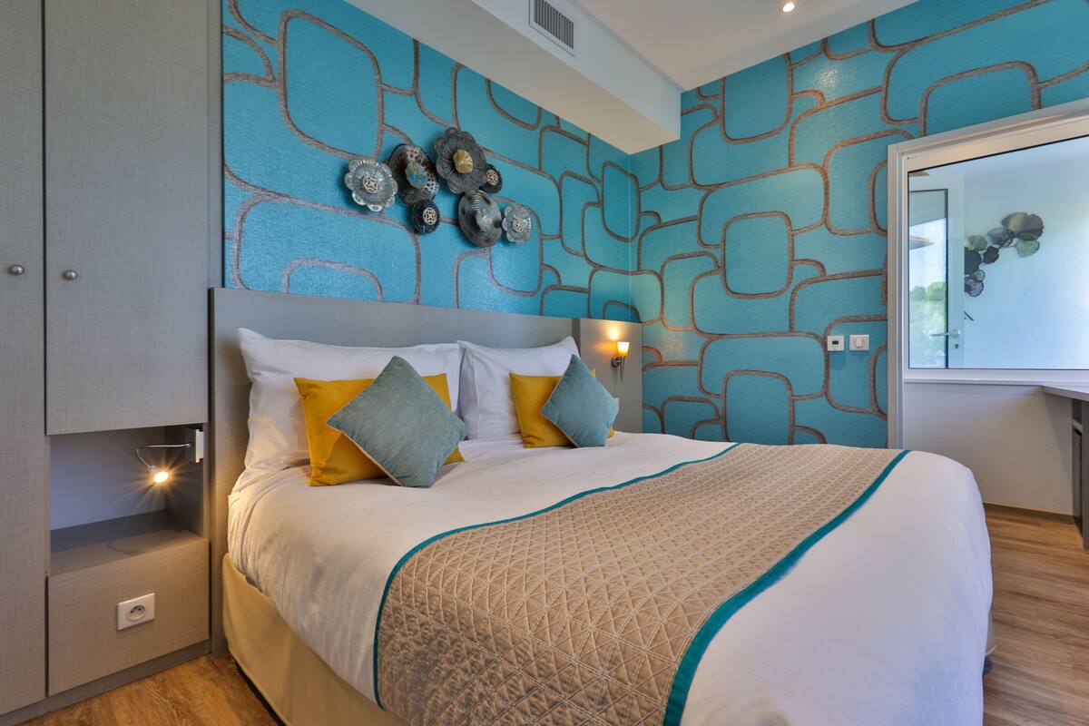 A modern bedroom with a geometric patterned wall, a bed with white and yellow bedding, and a nightstand with a lamp.