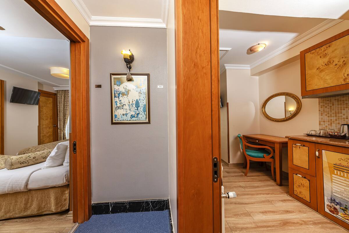 A hotel room with a bed, a desk, a chair, a mirror, and a small kitchenette.